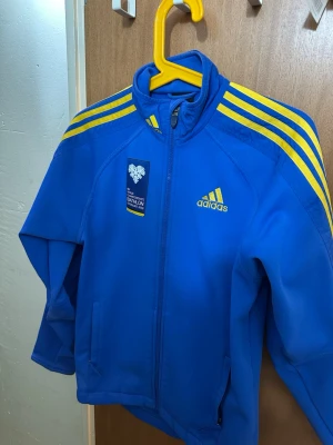 Blå Adidas träningsjacka med gula ränder - Snygg blå träningsjacka från Adidas med klassiska gula ränder på axlarna och dragkedja framtill. Jackan har hög krage, två sidofickor och ett tryckt märke på bröstet. Perfekt för sport eller uppvärmning.