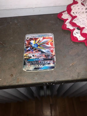 Pokémonkort Solgaleo GX - Säljer ett Pokémonkort med Solgaleo GX-motiv. Kortet har färgstark design med Solgaleo i silver, gult och blått på framsidan och klassisk blå baksida med Pokéball. Perfekt för samlare eller Pokémonfans som vill utöka sin kortsamling. Kortet är fejk