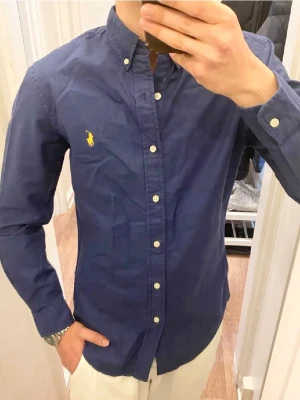 Mörkblå Skjorta från Ralph Lauren (Slim Fit) - Klassisk blå skjorta från Polo Ralph Lauren med button-down krage och den ikoniska gula logon broderad på bröstet. Skjortan har lång ärm, vita knappar framtill och är tillverkad i bomull. Perfekt för en clean och stilren look. Storlek S. Snabb Leverans!