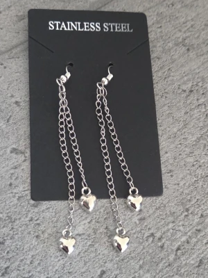 Silvriga örhängen med hjärtan - Säljer ett par långa silvriga örhängen med dubbla kedjor och små hjärtformade hängen längst ner. Örhängena har en modern och stilren look med blank finish. Perfekta för dig som gillar detaljer och vill ha något extra till din outfit. Krokar och kedjor i rostfritt stål (tål vatten och missfärgas inte). Ca 6,5 cm långa. 