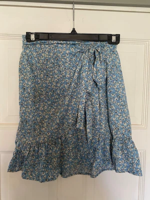 Blommig blå omlottkjol med volang - Söt blå omlottkjol med blommigt mönster och volangkant. Kjolen har knytband i midjan och är perfekt för varma dagar. Lätt och luftig modell som ger en avslappnad och trendig look.