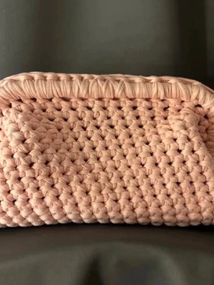 Rosa virkad kuvertväska - Handgjord kuvertväska i virkad design med mjukt, tjockt garn i en ljusrosa färg. Väskan har en rektangulär form och stängs med ett vikt lock. Perfekt accessoar för dig som gillar unika detaljer och trendig stil. Missonliknande