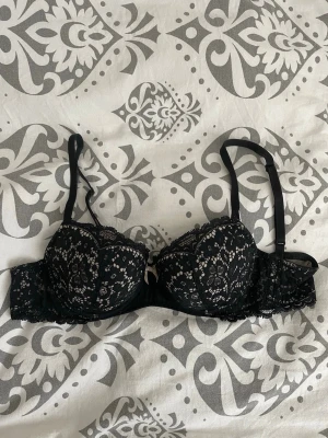 Svart spets-bh från Hunkemöller - Snygg svart bh från Hunkemöller med vadderade kupor och vacker spetsdetalj över hela bh:n. Justerbara axelband och liten rosett framtill. 