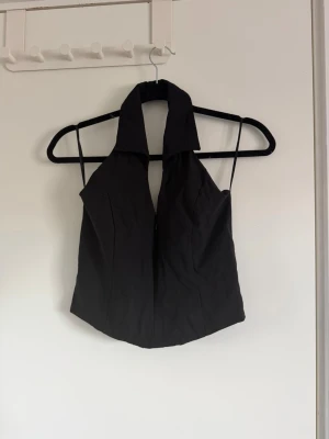 Svart halterneck korsettopp - Snygg svart korsettopp med halterneck och krage. Toppen har en djup v-ringning framtill och smala axelband. Perfekt för dig som vill ha en trendig och stilren look. Materialet känns slätt och lätt, troligen polyester.