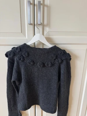 Mörkgrå stickad kofta med blommor - Mysig mörkgrå stickad kofta med knappar framtill och dekorativa virkade blommor längs axlar och ärmslut. Koftan har lång ärm och en något kortare, rak passform. 