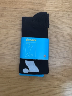Svarta fotbollsstrumpor från Seger - Säljer ett par svarta fotbollsstrumpor från Seger med vit detalj. Strumporna är knähöga och tillverkade i en mix av polyester och bomull, perfekta för dig som vill ha bekväma och funktionella strumpor till träning eller match.