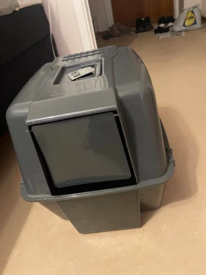 Grå kattlåda med lock och lucka - Säljer en grå kattlåda i plast med lock och svänglucka framtill. Lådan har handtag på ovansidan för enkel transport och ventilationshål. Perfekt för att hålla sanden inne och minska lukt i hemmet.