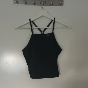 Svart träningslinne från Stronger - Snyggt svart träningslinne från Stronger med smala axelband som korsas i ryggen. Toppen har en stilren look och är tillverkad i ett stretchigt material som passar perfekt till träning. Enkel logga framtill och croppad passform. Aldrig använd. 