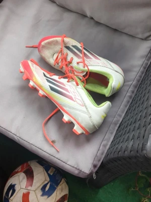 Adidas vita fotbollsskor med röda detaljer - Säljer ett par Adidas fotbollsskor i vitt med röda och svarta detaljer samt röda snören. Skorna har fasta dobbar och en låg profil, perfekt för snabba rörelser på planen. Insidan är limegrön för extra stilpoäng. Klassisk Adidas-design med tre svarta ränder på sidan.