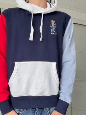 Flerfärgad hoodie från Polo Ralph Lauren - Snygg hoodie från Polo Ralph Lauren med marinblå kropp, vit huva och ficka, röd ärm och ljusblå ärm. Klassisk björnlogga broderad på bröstet. Mjuk bomullsblandning och stor känguruficka framtill. Perfekt för en avslappnad och trendig look.