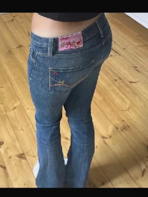 Blå bootcut jeans med broderi - Säljer ett par blå bootcut jeans med låg midja och snygga broderade detaljer på bakfickan samt en rosa etikett med blommor. Jeansen har klassisk femficksmodell och kontrastsömmar. Perfekta för dig som gillar en retro vibe!