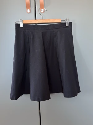 Svart kortkjol från Monki - Säljer en svart kortkjol från Monki i klassisk A-linjeform. Kjolen har en enkel och stilren design med diskreta sömmar framtill och är tillverkad i ett mjukt tyg som känns bekvämt. Perfekt att matcha med både sneakers och boots.