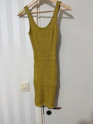 Ribbad senapsgul ärmlös klänning - Säljer en ribbad, figurnära senapsgul klänning med djup urringning både fram och bak. Klänningen är ärmlös och har en kort längd som passar perfekt till varmare dagar. Materialet är stretchigt och bekvämt, vilket ger en snygg passform.
