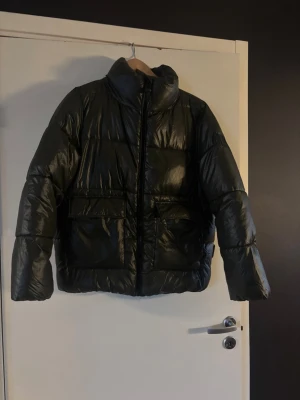 Mörkgrön pufferjacka från Navahoo - Snygg mörkgrön pufferjacka från Navahoo med hög krage och två stora fickor framtill. Jackan har glansig yta och dragkedja hela vägen upp. Perfekt för kalla dagar och riktigt skön att bära. Materialet är syntet och jackan har en modern, boxig passform.