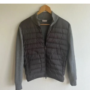 Grå Moncler cardigan - Eftertraktade cardiganen från Moncler med quiltad framsida och stickade ärmar. Jackan har en dragkedja framtill. Mycket bra skick, skriv vid flera bilder. Tyvärr inget kvitto men scan och tags finns!