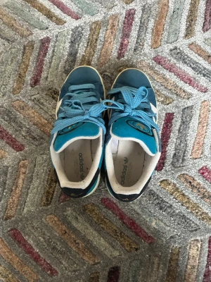Blå Adidas Originals sneakers - Snygga blå Adidas Originals sneakers med klassiska vita ränder på sidan och guldfärgad logga på tungan och hälen. Skorna har blå skosnören, vit sula och ovandel i mocka och textil. Perfekta för dig som gillar retrostil och streetwear.