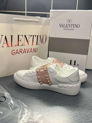 Valentino Garavani vita sneakers - Snygga vita sneakers från Valentino Garavani med ljusrosa band och ikoniska nitar i gummi längs sidorna. Skorna har en klassisk låg siluett, vita snören och detaljer i läder. Perfekta för dig som vill ha en exklusiv och trendig look.