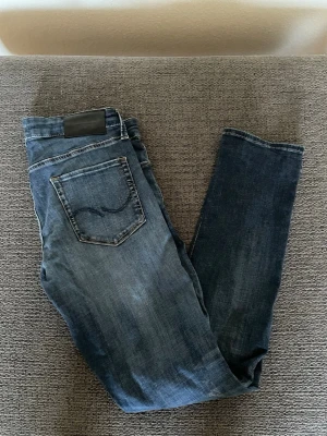 Jack and Jones jeans  - Snygga blå slim fit jeans från Jack and Jones  med klassisk femficksdesign och diskret vågdetalj på bakfickan. Jeansen har en mörk tvätt och är tillverkade i mjuk bomullsdenim. Perfekta för en avslappnad och trendig look.