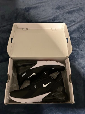 Nike Air Max 270 svarta sneakers - Snygga Nike Air Max 270 sneakers i svart med vita detaljer och logga. Skorna har en luftig mesh-överdel, stor synlig Air-enhet i hälen och vit sula. Perfekta för dig som gillar sportig och modern stil. Klassisk Nike-design med bekväm passform.