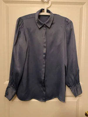 Blå satinblus med puffärm - Snygg blå blus i glansigt satintyg med klassisk krage och puffiga axlar. Långa ärmar med breda manschetter och knappar vid ärmslut. Perfekt för dig som vill ha en stilren men ändå trendig look. Kolla gärna mina andra annonser❗️