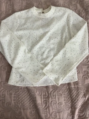 Vit prickig blus med lång ärm - Snygg vit blus med små svarta prickar och lång ärm. Blusen har en hög halsringning och knäppning bak i nacken med två pärlknappar. Tillverkad i ett lätt, något transparent tyg som ger en luftig känsla.