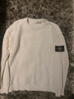 Vit stickad tröja från Stone Island - Vit stickad tröja från Stone Island med klassisk rund halsringning och ribbade muddar. Tröjan har den ikoniska Stone Island-loggan på vänster ärm. Perfekt för dig som gillar stilrena och exklusiva plagg.