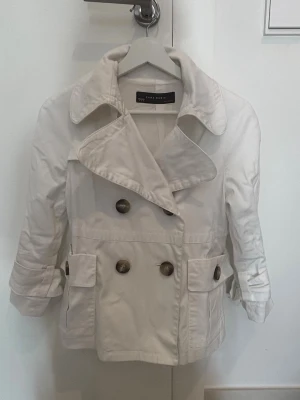 Vit trenchcoat från Zara Basic - Kort vit trenchcoat från Zara Basic med dubbelknäppning och stora bruna knappar. Jackan har bred krage, två fickor med lock och justerbara band vid ärmsluten. Perfekt för dig som gillar klassisk och stilren look.