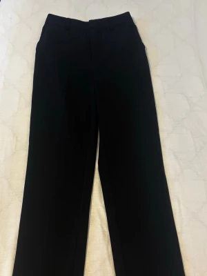 Svarta raka kostymbyxor - Snygga svarta kostymbyxor med rak passform och klassiska bälteshällor. Byxorna har pressveck framtill och är perfekta för en stilren look. Materialet känns mjukt och följsamt, men exakt tyg kan ej avgöras. Från cubus 