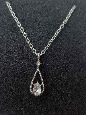 Silverfärgat halsband med droppformad kristall - Snyggt halsband i silverfärg med en tunn kedja och ett droppformat hänge. Hänget är dekorerat med små gnistrande stenar och en större klar kristall i mitten. Perfekt för dig som gillar stilrena och eleganta smycken. Materialet ser ut att vara metall med glittrande stenar.