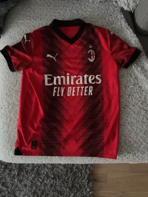  Puma  - Snygg AC Milan matchtröja från Puma i rött och svart med klubbmärke och Puma-logga på bröstet. Tryck med 'Emirates Fly Better' på framsidan och 'wefox' på ryggen. Kortärmad med svart krage och detaljer. Perfekt för fotbollsträning eller att bära till vardags. 2023/24 stället
