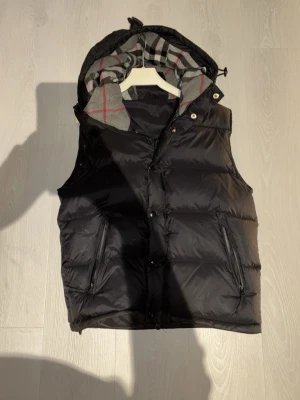 Svart pufferjacka från Burrybery - Svart pufferjacka med stor huva och rutig grå, vit och röd insida. Jackan har dragkedja, tryckknappar och två sidofickor med dragkedja. Märke 'BURRYBERY LONDON ENGLAND' på bröstet. Perfekt för kalla dagar med sin tjocka och quiltade design.