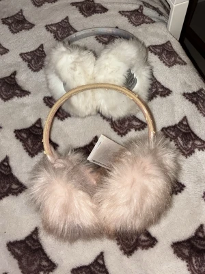Säljer Två fluffiga öronmuffar i pastell - De kostar 120kr tillsammans! Säljer två par öronmuffar, ett par i ljusrosa med glittrigt band och ett par i vitt med silverdetalj. Båda har mjuk, fluffig fuskpäls och är perfekta för kalla dagar. Passar dig som vill hålla värmen med stil.