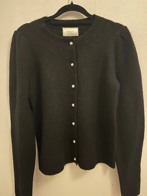 Svart kofta med pärlknappar Neo Noir - Stilren svart kofta från Neo Noir med ribbstickad struktur och dekorativa pärlknappar framtill. Rund halsringning och puffade axlar ger en trendig touch. Perfekt att slänga över en topp för en snygg lager-på-lager-look.