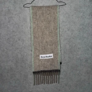 Beige halsduk från Acne Studios - Snygg beige halsduk från Acne Studios med ljusgröna kanter och klassiska fransar. Tillverkad i mjukt material som känns lyxigt och har en tydlig logotyp framtill. Perfekt accessoar för att lyfta din outfit.