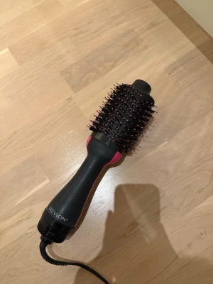 Revlon varmluftsborste svart/rosa - En elektrisk varmluftsborste från Revlon i svart och rosa med ovala borsthuvud och piggar med rosa toppar. Handtaget är ergonomiskt och borsten har sladd med vanlig stickkontakt. Perfekt för att styla och torka håret samtidigt.