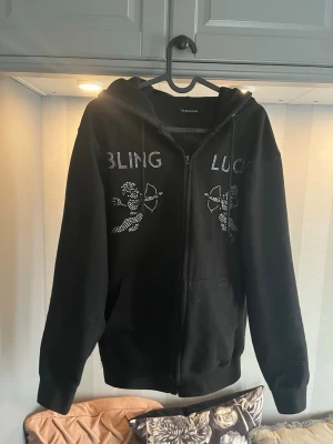 Svart hoodie från bling Lucid - Svart hoodie med dragkedja från Bling Lucid. Framsidan har glittrande text och motiv av två änglar i strass. Hoodien har huva och fickor framtill. Perfekt för dig som gillar streetwear och vill sticka ut med detaljer.