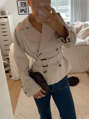 Kort beige trenchcoat med bälte - Stilren beige trenchcoat med bred krage, dubbelknäppning och matchande bälte. Köpt för ungefär en månad sedan men kommer tyvärr inte till användning🥲❤️