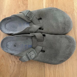 Birkenstock Boston mockatofflor grå - Säljer ett par klassiska Birkenstock Boston tofflor i grå mocka. De har justerbart spänne i metall och en bekväm fotbädd. Yttersulan är i gummi med vågigt mönster för bra grepp. Perfekta för dig som gillar stilrena och tidlösa skor.