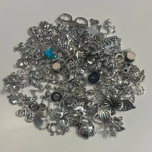 80st mixade silverfärgade berlocker - Stort blandat paket med ca 80st silverfärgade berlocker i olika former. Perfekt för att skapa egna smycken eller dekorera accessoarer. Materialet är huvudsakligen metall.