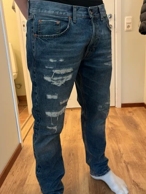Blå slitna jeans från Dondup - Säljer ett par blå jeans från Dondup med slitna detaljer och distressed look framtill. Klassisk femficksmodell med raka ben och normal midja. Jeansen har en snygg tvättad finish och är tillverkade i denim.