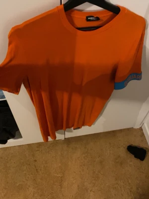 Orange t-shirt från Dsquared2 - Snygg orange t-shirt från Dsquared2 med blå detalj och logga på ärmen. Klassisk rund hals och korta ärmar. Perfekt för dig som vill sticka ut med färg och stil. Tillverkad i mjuk bomull för skön känsla hela dagen.