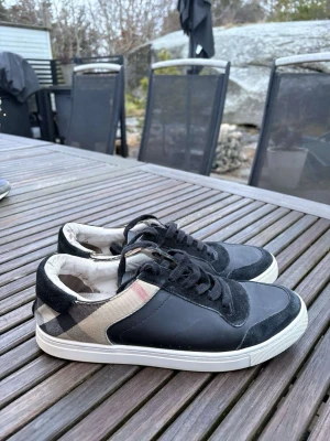 Burberry sneakers med rutigt mönster - Snygga sneakers från Burberry med klassiskt beige, svart och rött rutmönster på sidan. Skorna har svarta mockadetaljer, svarta skosnören och vit sula. Perfekta för dig som vill ha en stilren och trendig look med lyxig känsla.