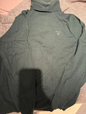 Mörkgrön polotröja från Gant - Mörkgrön stickad polotröja från Gant med diskret logga på bröstet. Tröjan har lång ärm, ribbade muddar och hög polokrage. Perfekt för lager på lager och en clean look. Materialet känns mjukt och följsamt.