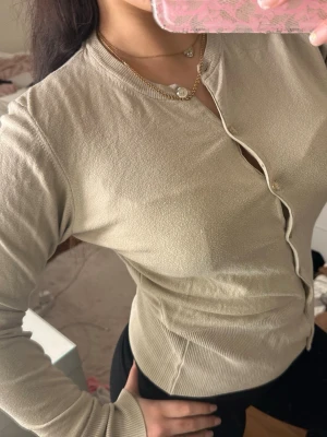 Beige ribbad cardigan med knappar - Snygg beige cardigan med ribbade muddar och knäppning framtill. Figurnära passform och rund halsringning. Perfekt att styla med jeans eller kjol för en clean look.