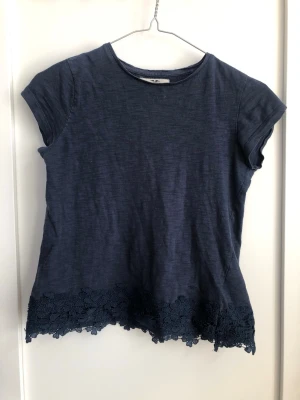 Mörkblå topp med spetskant - Snygg mörkblå topp med korta ärmar och rund halsringning. Nedtill finns en dekorativ spetskant med blommönster som ger en extra detalj. Perfekt att matcha med jeans eller kjol för en trendig look.