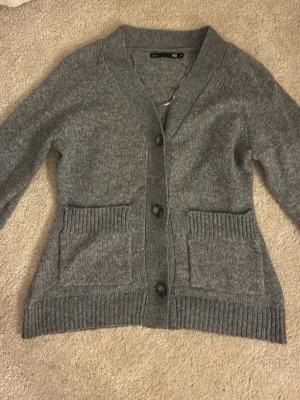 Grå kofta med fickor och knappar - Mysig grå kofta med v-ringning och tre stora knappar framtill. Två rymliga fickor och ribbade muddar vid ärmslut och nederkant. Den kommer nt till användning, har använt 1 gång. 