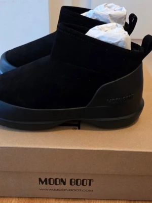 Svarta Moon Boot ankelboots - Stilrena svarta Moon Boot ankelboots med låg profil och robust sula. Ovandelen är i mockaliknande material och sulan i slitstark gummi. Diskret logga på sidan och praktisk hälflik för enkel påtagning. Perfekta för kalla dagar när du vill hålla stilen.