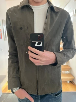 Olivgrön overshirt från J.Lindeberg - Snygg olivgrön overshirt från J.Lindeberg med dragkedja framtill och en bröstficka med knapp. Overshirten har klassisk krage och är tillverkad i ett tunt, mjukt bomullsliknande material. Perfekt att bära över en t-shirt för en avslappnad look. Lite solblekt på vissa ställen men gör att det ser stilrent ut!
