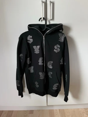 Svart hoodie med blingade valutasymboler - Snygg svart hoodie med dragkedja och huva, täckt av blingade valutasymboler som dollar, euro och pund i silverfärgade stenar. Hoodien har kängurufickor och ribbade muddar vid ärmar och nederkant. Perfekt för dig som gillar statement-plagg.