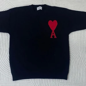 Svart stickad tröja från Ami Paris - Säljer en svart stickad tröja från Ami Paris med deras ikoniska röda hjärta och bokstaven A broderat på bröstet. Tröjan har rund halsringning och ribbade muddar. Perfekt för dig som gillar stilrena plagg med en twist. Storlek M men passar mer S